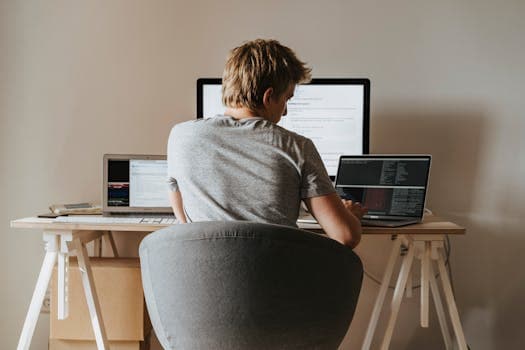 Young man coding at home office with multiple laptops, representing remote work and technology.