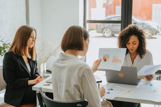 Three women engaged in a business meeting, discussing charts and data in a modern office setting.