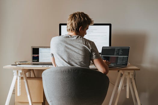 Young man coding at home office with multiple laptops, representing remote work and technology.
