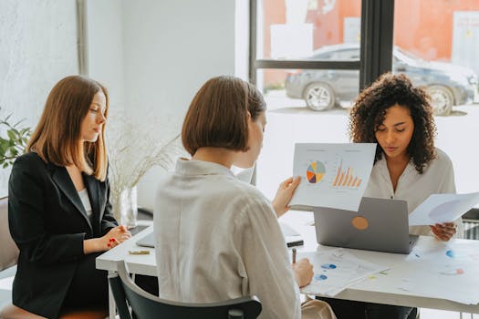 Three women engaged in a business meeting, discussing charts and data in a modern office setting.